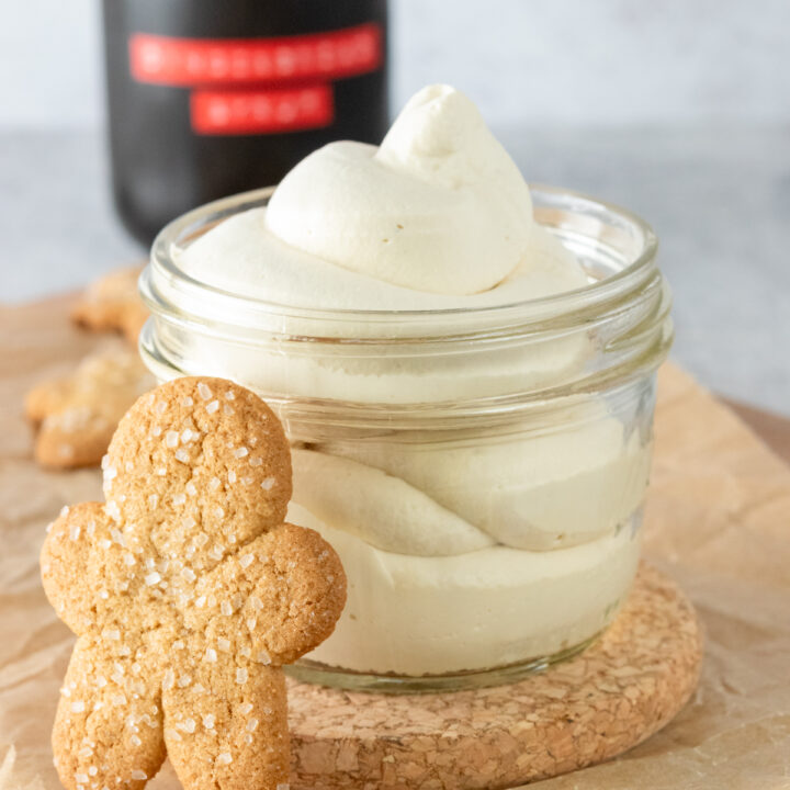 Homemade gingerbread whipped cream in front of a bottle of gingerbread syrup and with a gingerbread cookie leaning against jar of whipped cream.