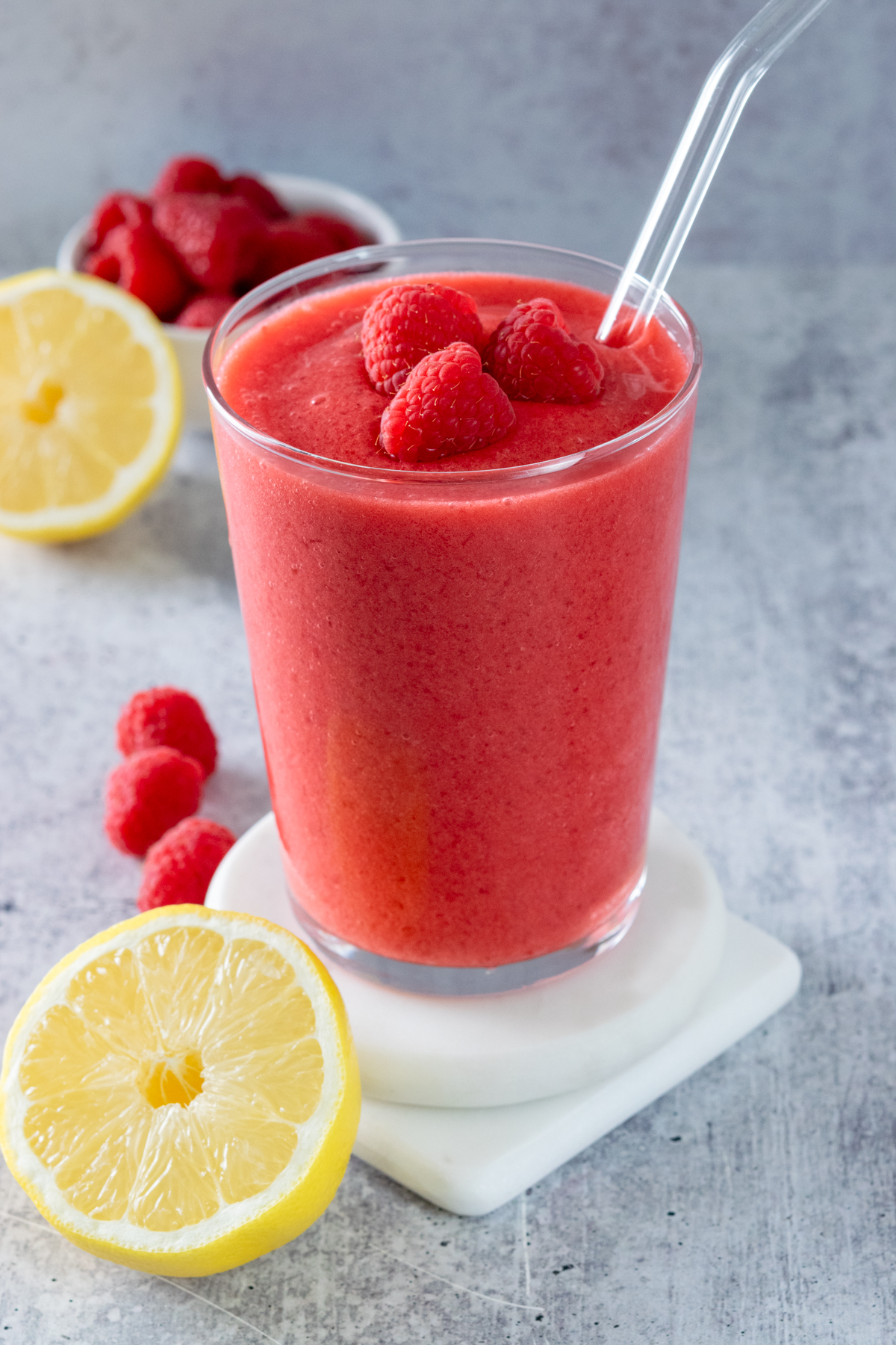 Frozen raspberry lemonade in a glass with straw with three raspberries on top. The drink is next to half a lemon and a few fresh raspberries.