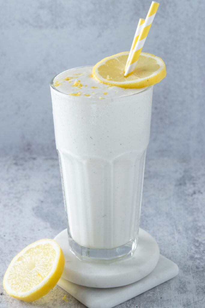 Creamy and slushy frosted lemonade drink in a tall glass with straws, lemon zest and a sliced lemon on the side.
