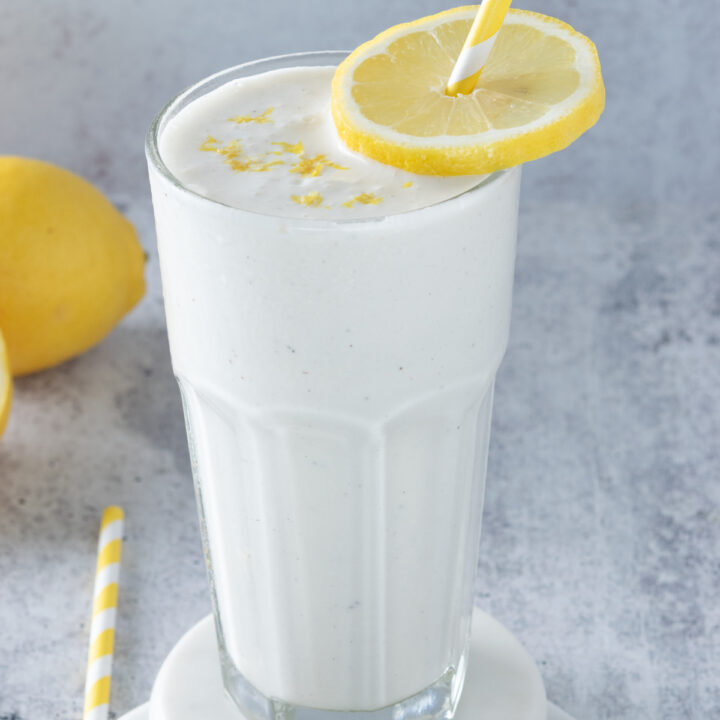 Frosted lemonade drink in a tall frosted glass, garnished with lemon slice and lemon zest. The cup has a yellow striped straw in it and is next to a lemon a a few more straws on the counter.