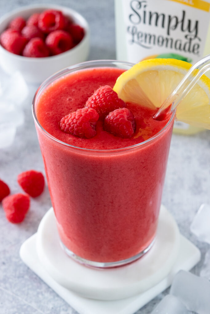 A frozen raspberry lemonade garnished with three raspberries and a lemon slice, sitting in front of a bottle of lemonade and bowl of rapsberries.