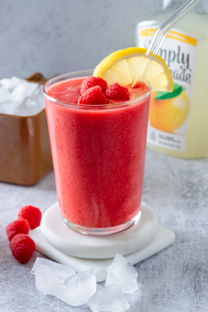 A blended raspberry lemonade topped with raspberries, next to a scoop of ice cubes and bottle of lemonade.