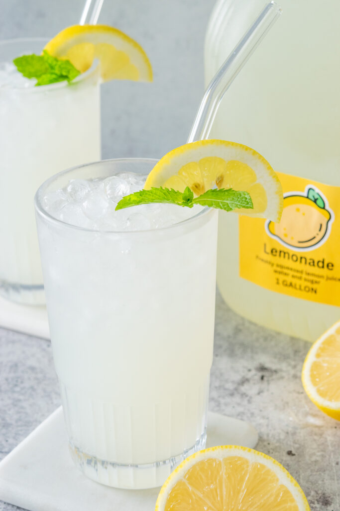 Two glasses of ice cold lemonade next to a gallon of homemade lemonade that has a yellow 1 Gallon Lemonade label on it.