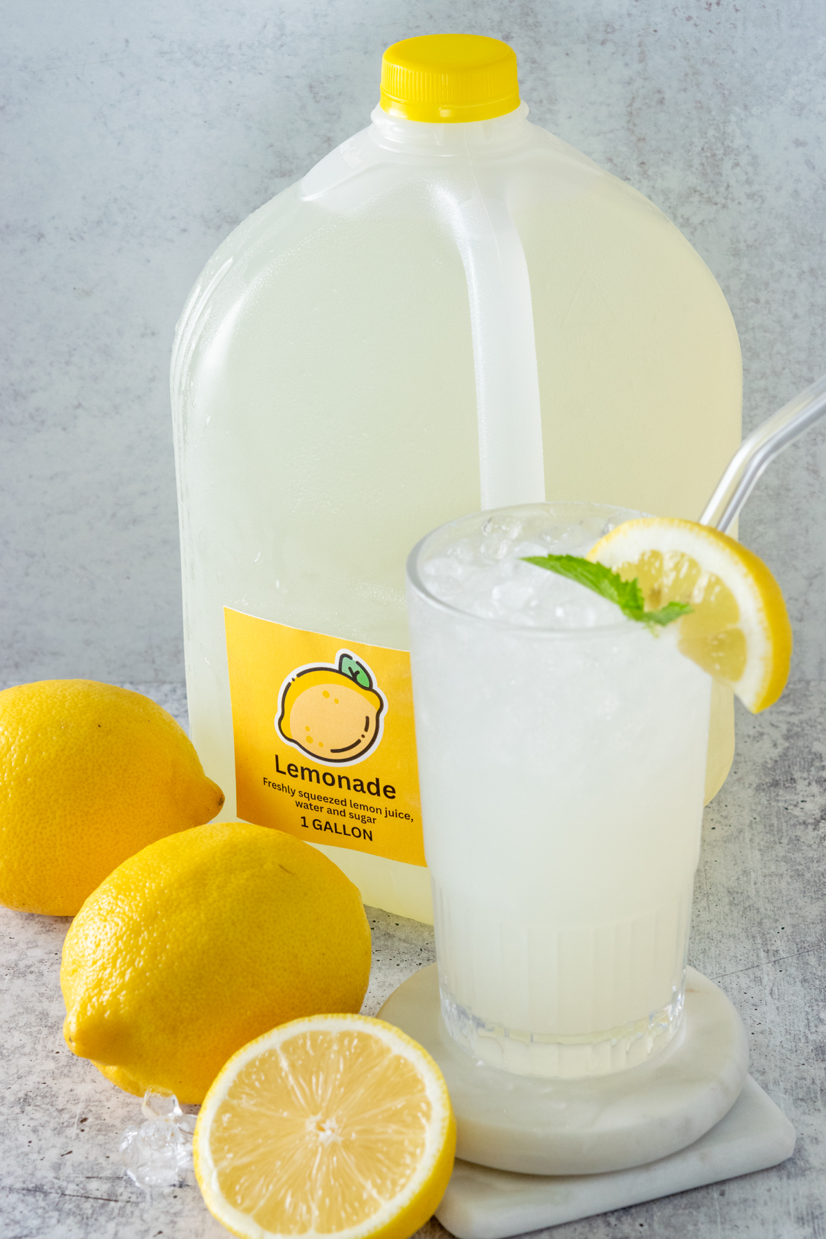 Glasses of ice cold lemonade next to three lemons and a gallon of homemade lemonade that has a yellow 1 Gallon Lemonade label on it.