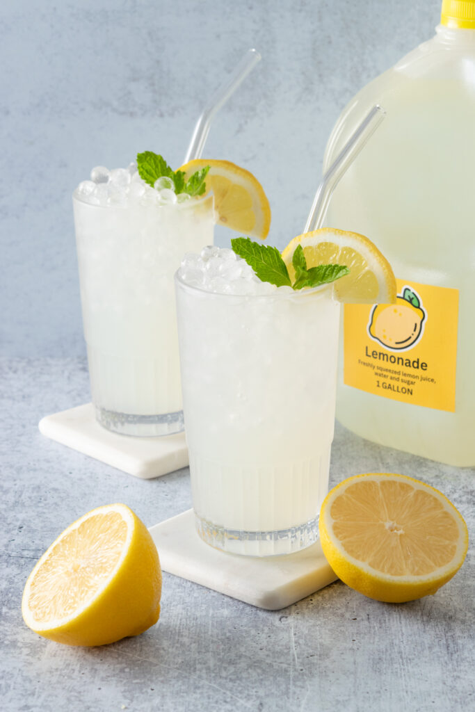 Two ice cold glasses of lemonade next to a gallon jug of homemade lemonade which has a yellow lemonade label on it.