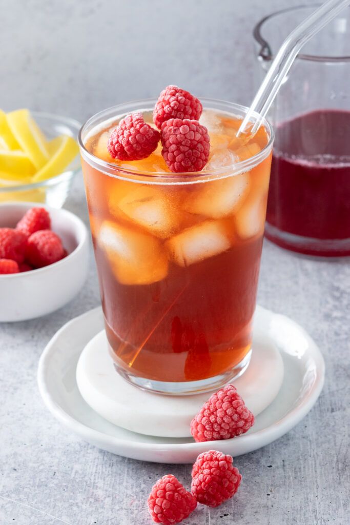 Glass of raspberry iced tea with lots of ice and raspberries in it, sitting in front of bowls of raspberries, lemon wedges and jar of homemade syrup.