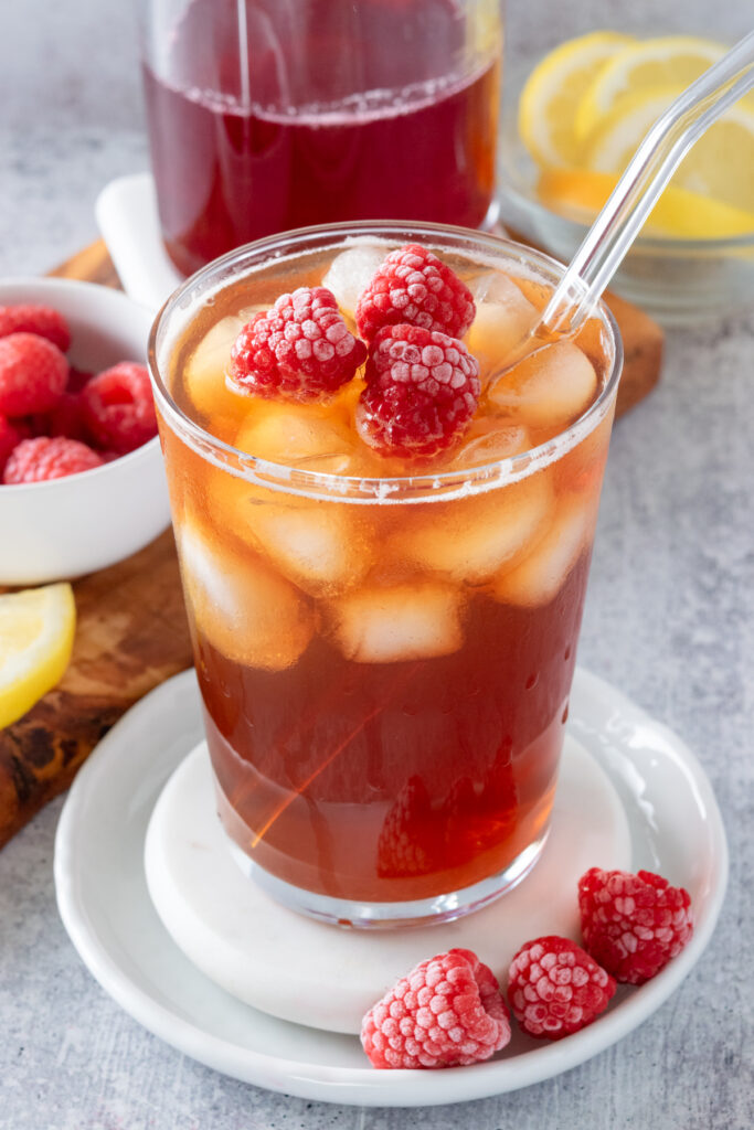 Ice cold raspberry tea drink with raspberries floating on top. The drink is sitting next to bottle of homemade raspberry syrup and bowl of raspberries and lemon wedges.