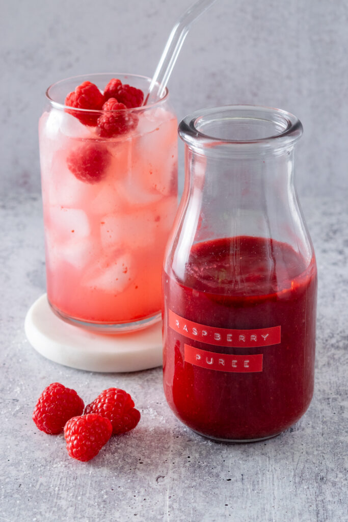Homemade raspberry puree in a small glass bottle with a raspberry puree label  on it. The puree is next to a raspberry lemonade drink and three raspberries.