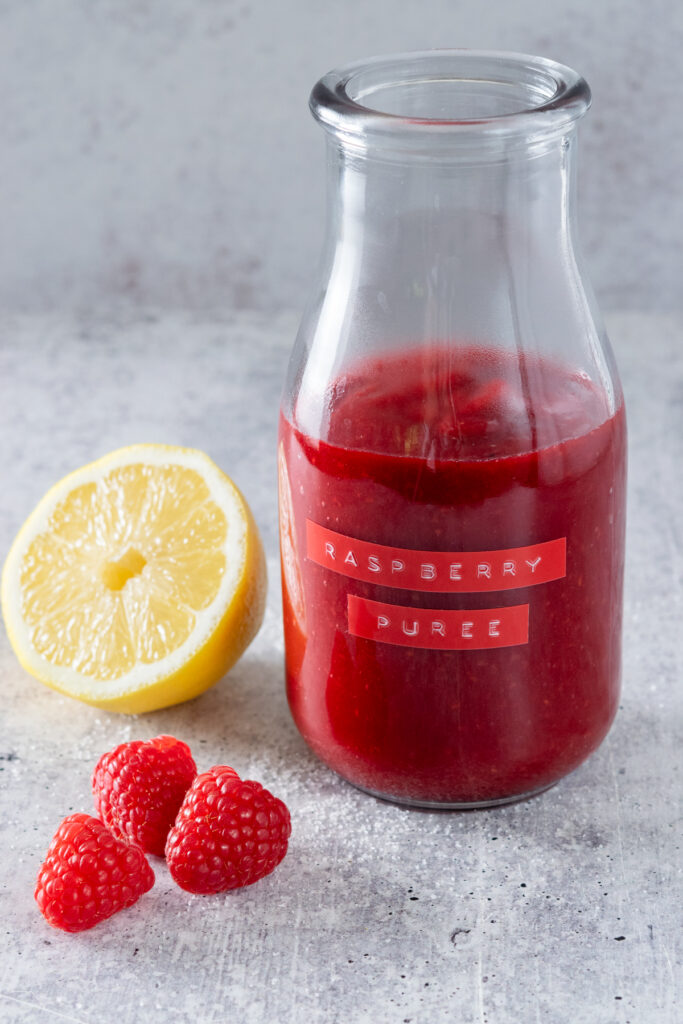Homemade raspberry puree in a bottle with raspberry puree label next to three raspberries and lemon wedge.