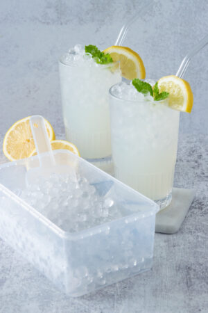 Container of round mini ice cubes and scoop next to two glasses of lemonade full of ice.