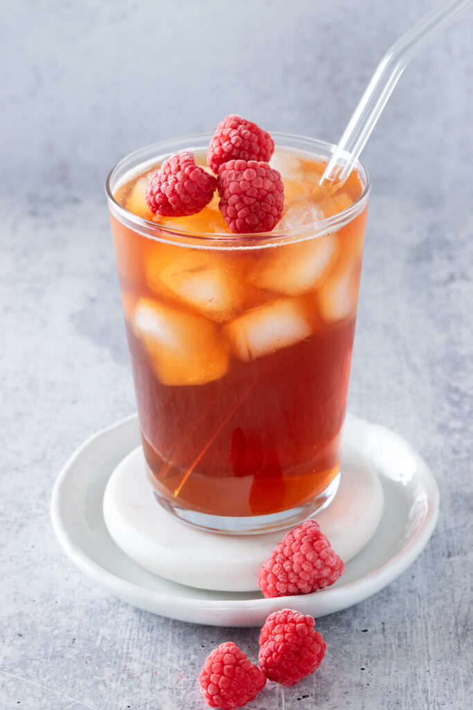 Refreshing glass of raspberry iced tea garnished with frozen raspberries, in a glass with glass straw on white marble coaster.