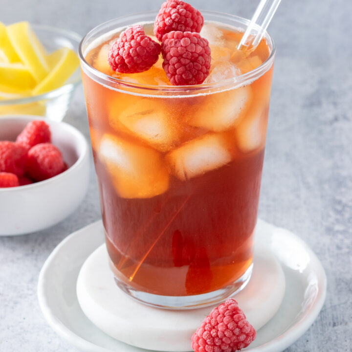 Glass of homemade raspberry iced tea with frozen raspberries on top. The drink is next to a small bowl of raspberries and lemon wedges.