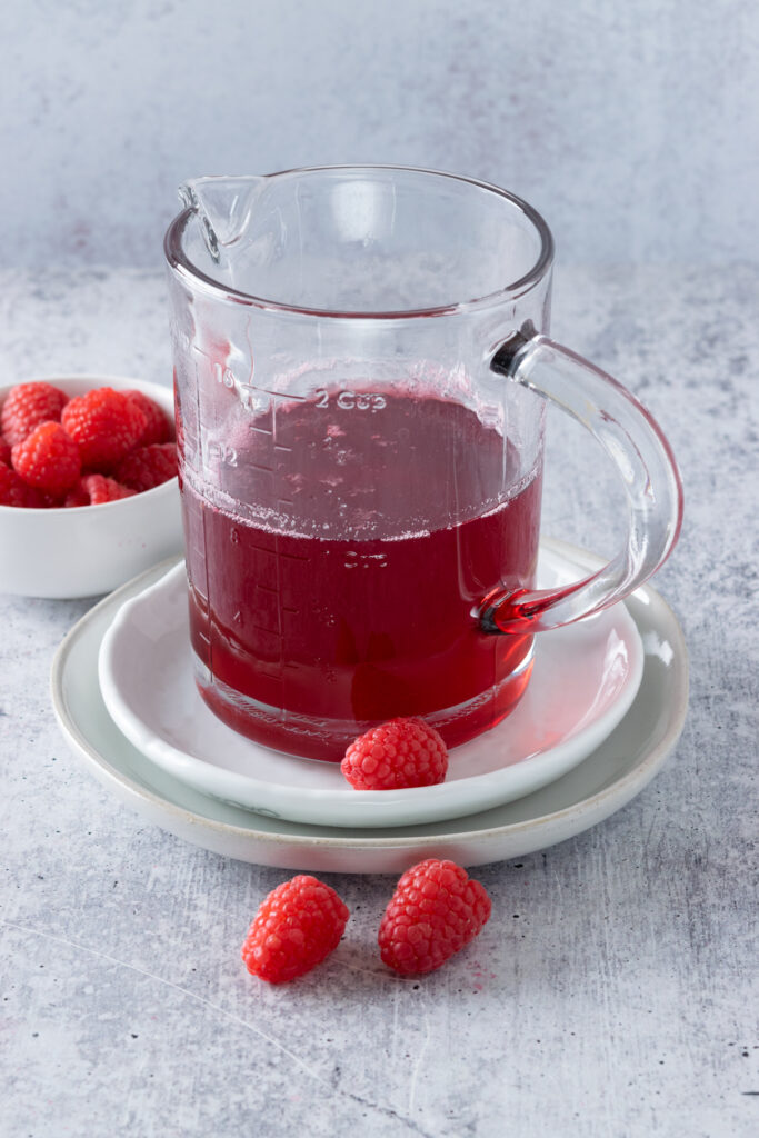 Jar of homemade raspberry syrup sitting on a plate with fresh raspberries next to it.