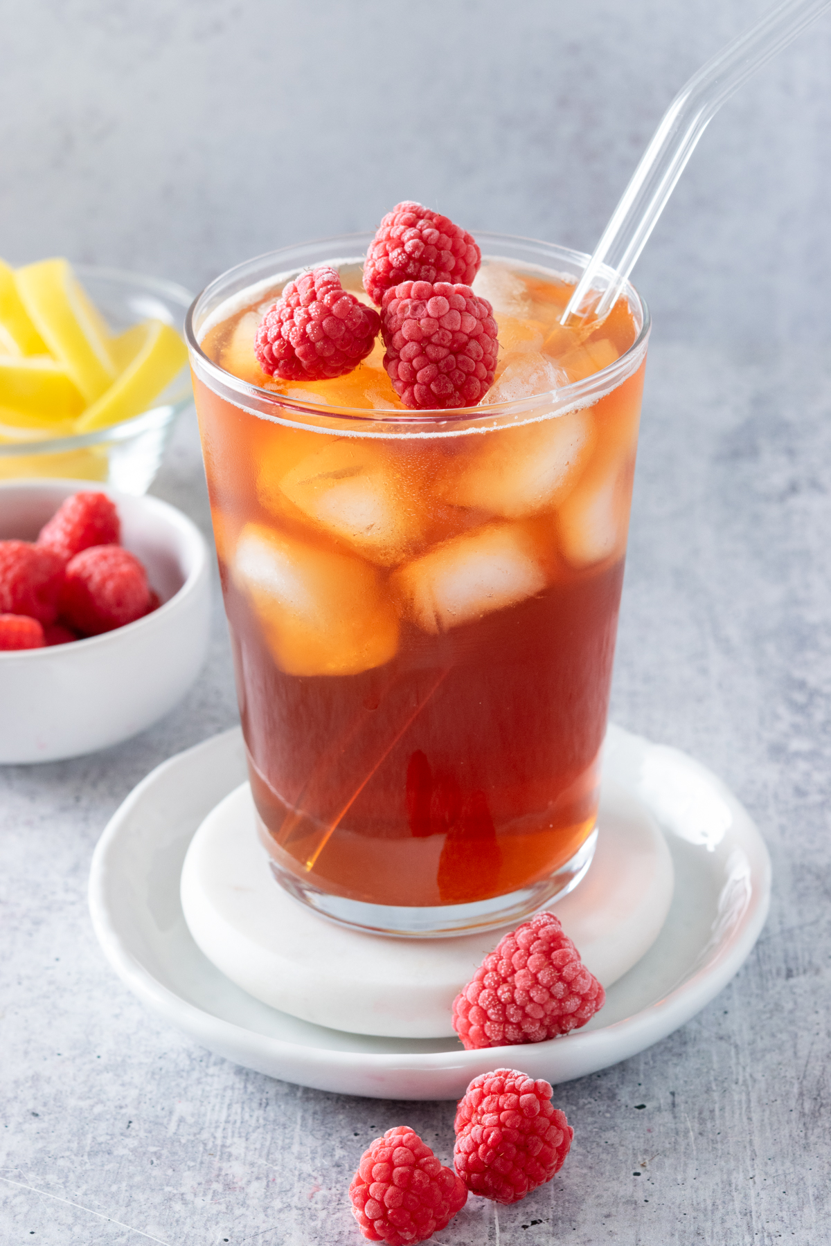 Glass of homemade raspberry iced tea with frozen raspberries on top. The drink is next to a small bowl of raspberries and lemon wedges.