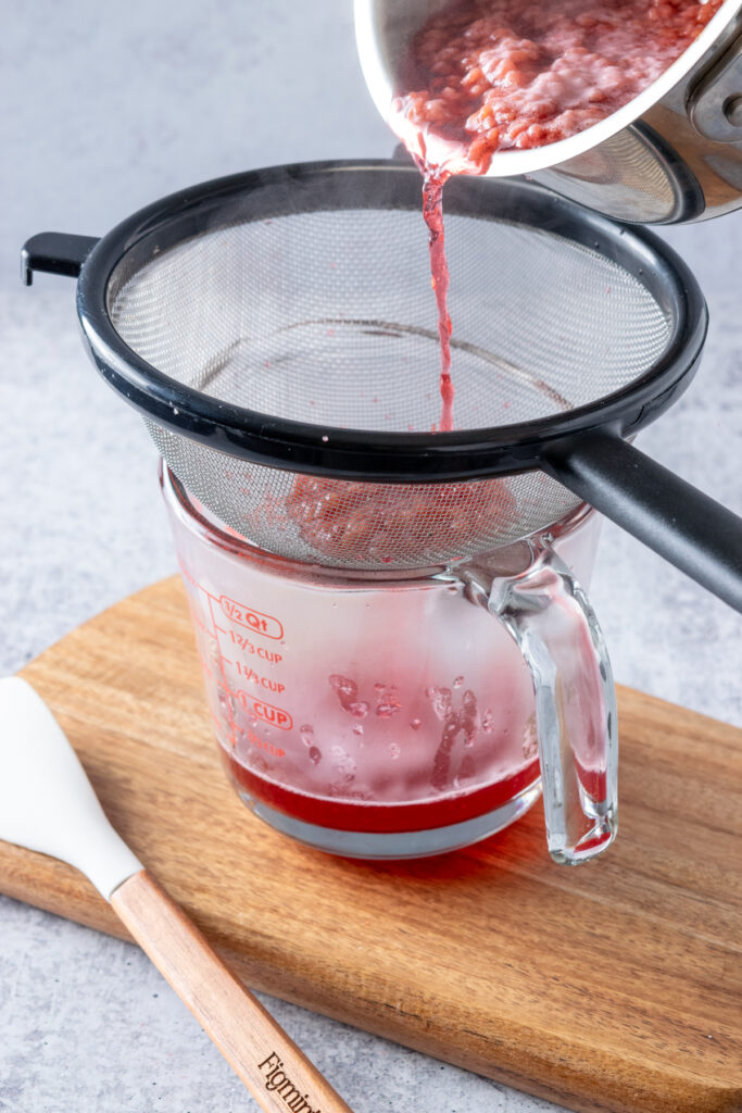 Pouring raspberry juice from the saucepan into a glass measuring cup through a strainer to catch the solids.