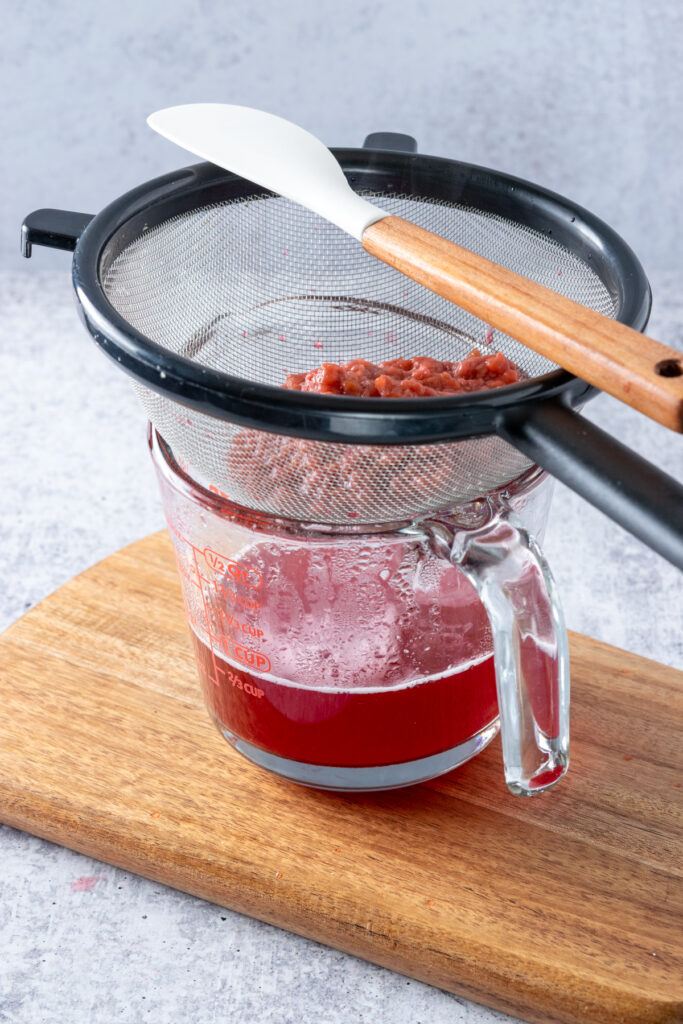 Homemade raspberry syrup in a glass measuring cup, with strainer full of pulp on top.