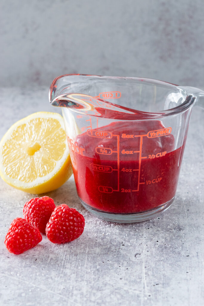 Glass measuring cup with homemade raspberry puree in it next to half a lemon and some fresh raspberries.