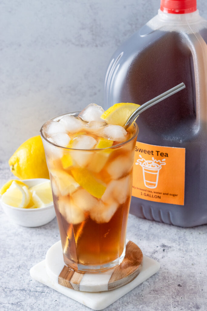 A full glass of iced tea with lemon in it, in front of a gallon container of homemade sweet tea.