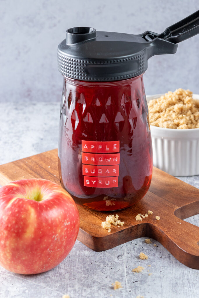 A fluted glass mason jar that has a spout lid on it, filled with homemade apple brown sugar syrup. The bottle is sitting on a wood board next to an apple and bowl of brown sugar.