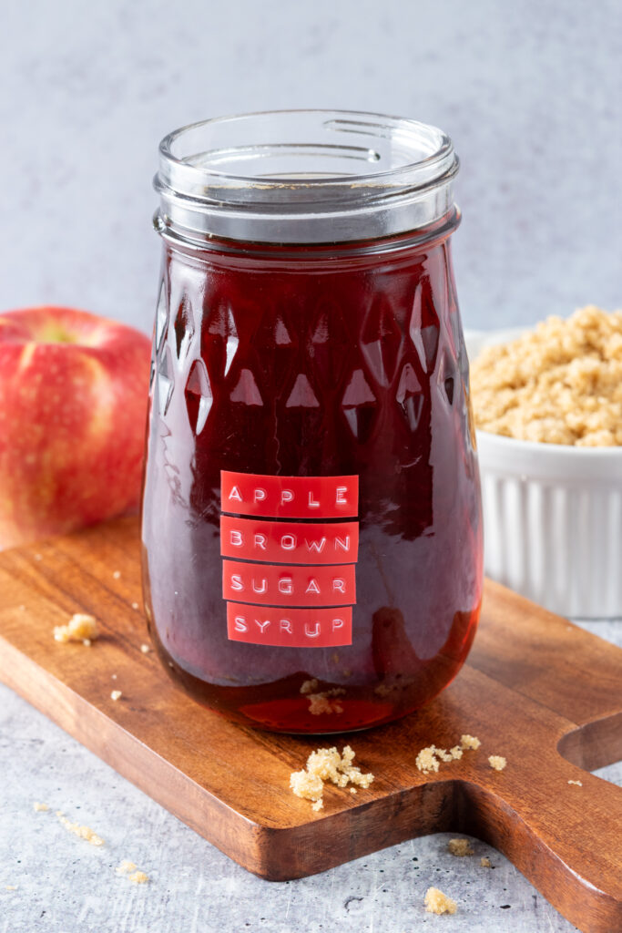 Jar of homemade apple brown sugar syrup next to a red apple and bowl of brown sugar.