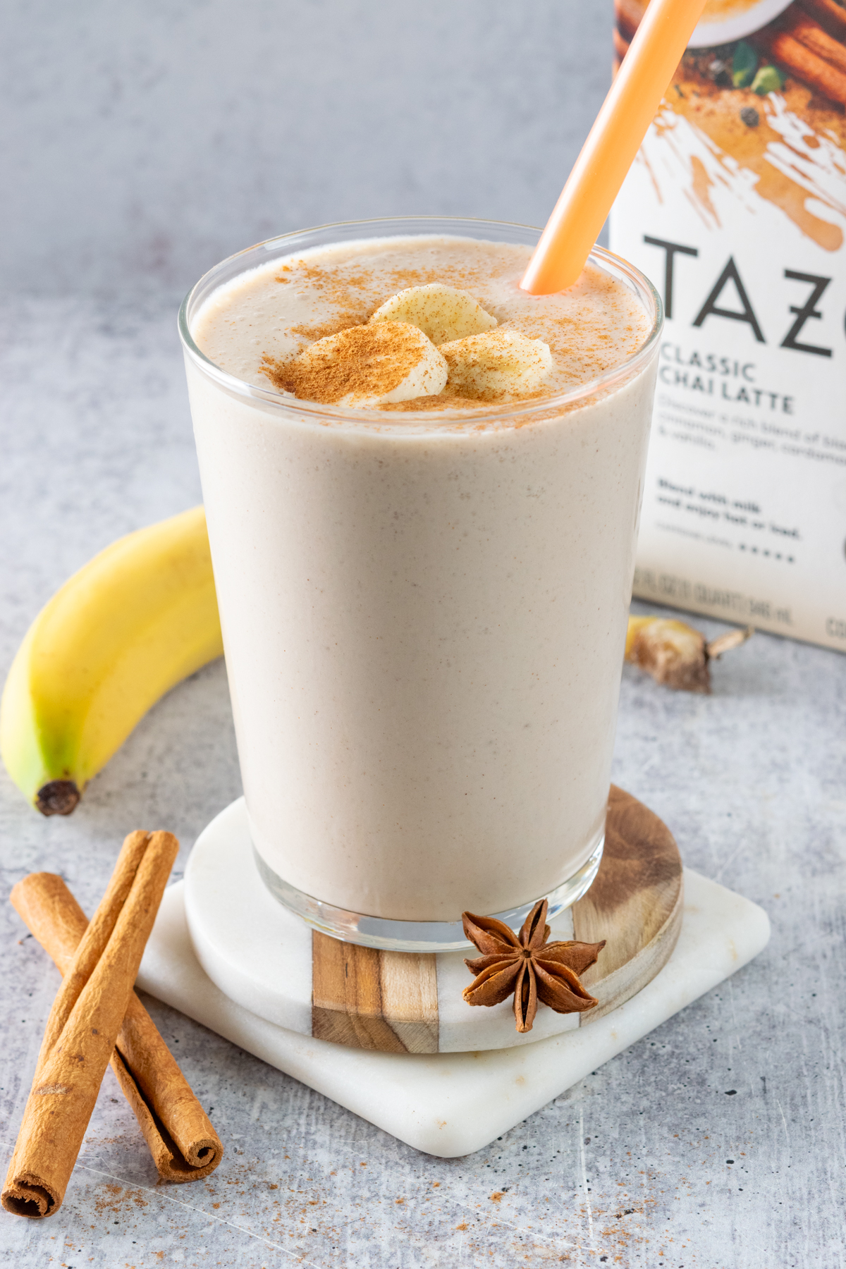 A homemade banana chai smoothie garnished with a few slices of banana and cinnamon, next to some cinnamon sticks, a banana and carton of chai tea latte concentrate.