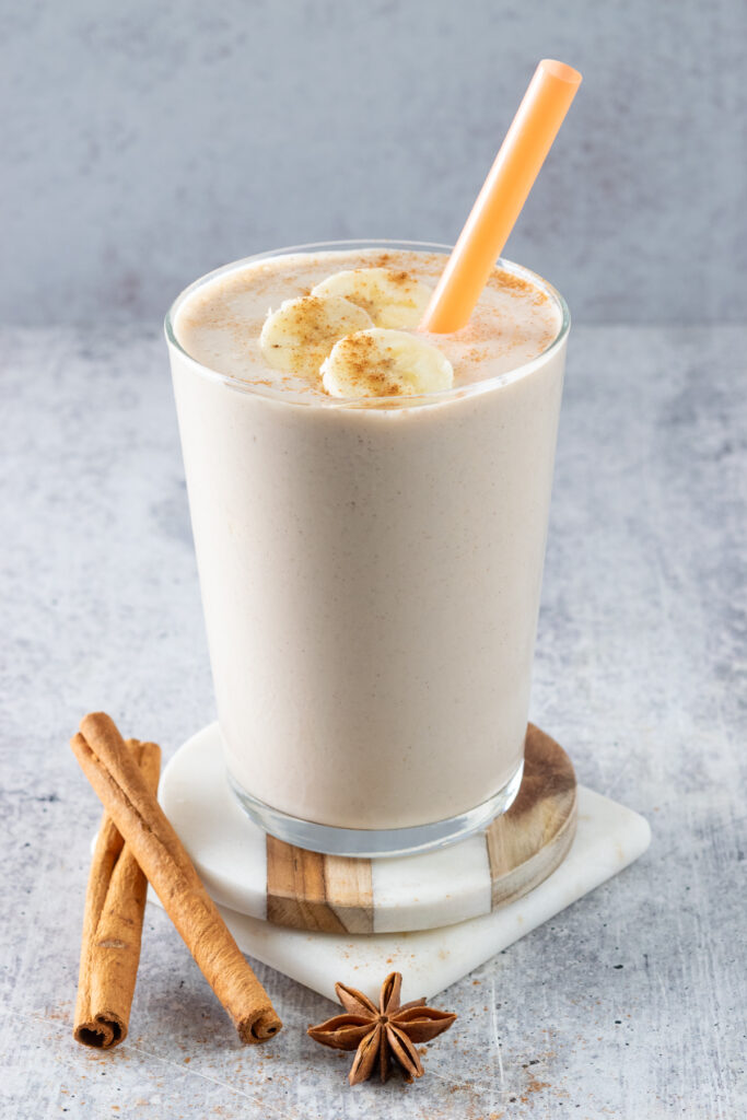 A chai banana smoothie that's topped with three slices of banana that have ground cinnamon sprinkled on top of them. The cup has an orange straw and is on a brown and white coaster which is next two a star anise and two cinnamon sticks.