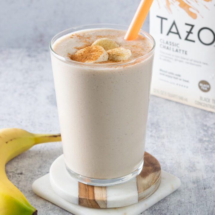 A banana chai smoothie topped with slices of banana and ground cinnamon, next to an unpeeled banana and carton of chai tea concentrate.