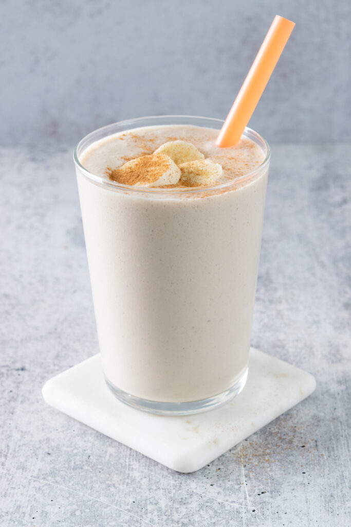 A banana chai smoothie garnished with slices of banana and ground cinnamon. The glass has an orange straw in it and is sitting on a white marble coaster.