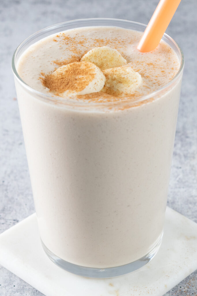 Banana chai smoothie in a glass cup with an orange straw and slices of banana on top with cinnamon sprinkled over the bananas.