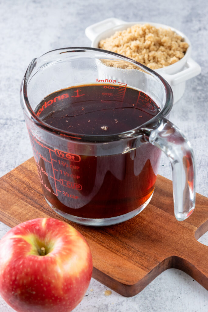 A 2-cup measuring cup full of the homemade apple brown sugar syrup that's sitting on a small wood cutting board next to a red apple and bowl of brown sugar.