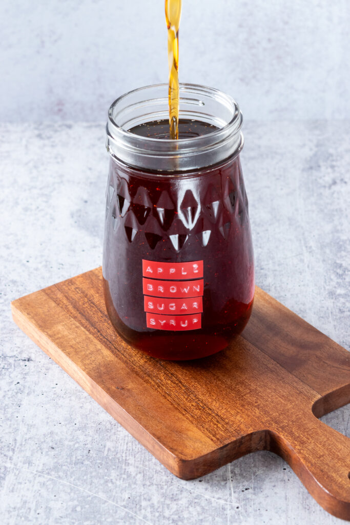 Stream of the syrup being transferred into a glass mason jar for storage, that has a red label on it that says, apple brown sugar syrup.