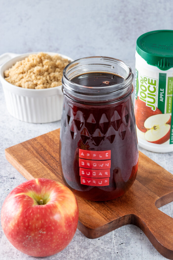 Open mason jar filled with homemade apple brown sugar syrup, next to a container of frozen apple juice concentrate, an apple and small bowl of brown sugar.