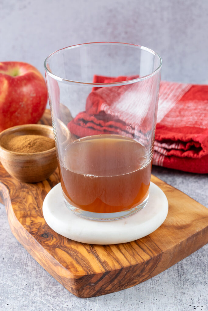 Chai concentrate poured partially filling a clear glass cup which is on a wood board and next to a small bowl of cinnamon, an apple and a red towel.