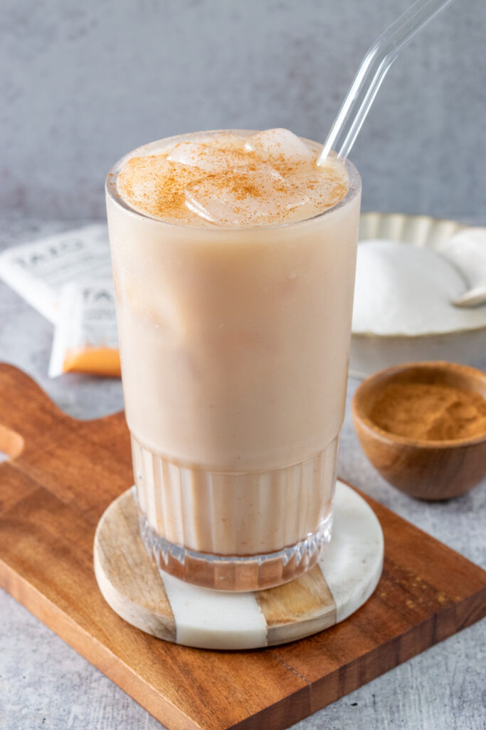 An iced chai tea latte in a tall glass filled with ice and cinnamon sprinkled on top, next to a small wood bowl of ground cinnamon and a couple tea bag wrappers in the background.