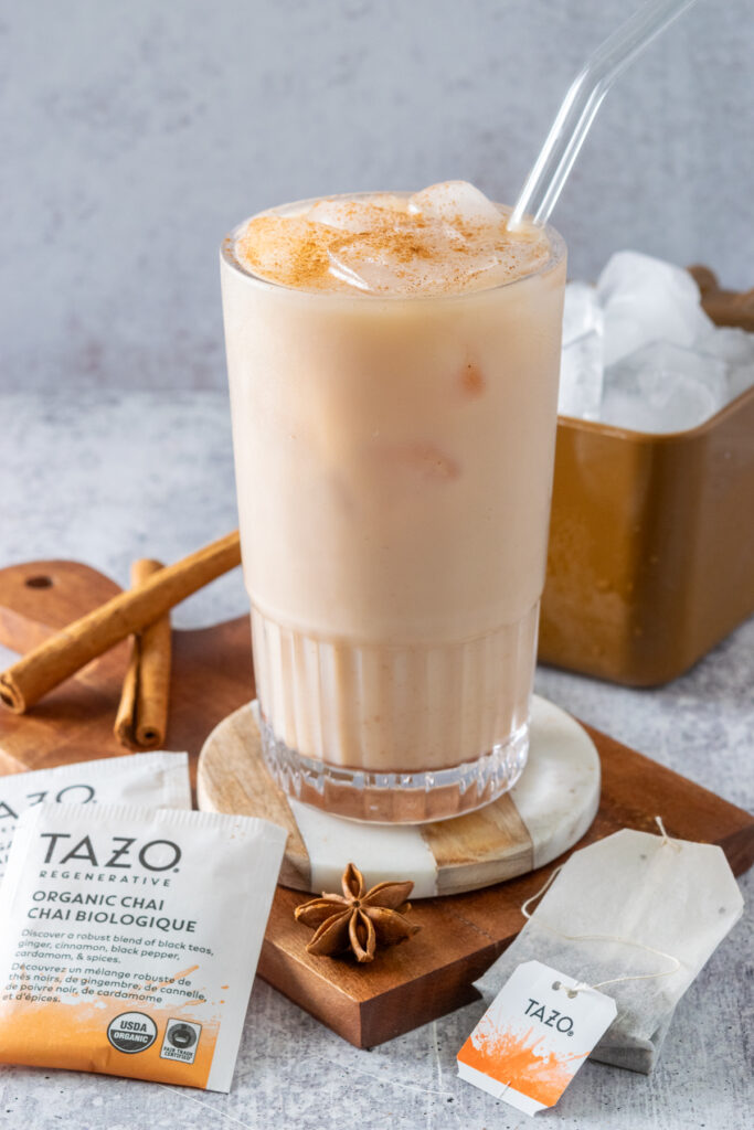 A homemade iced chai tea latte with cinnamon sprinkled on top, next to two cinnamon sticks, one star anise, three chai tea bags and a scoop of ice.