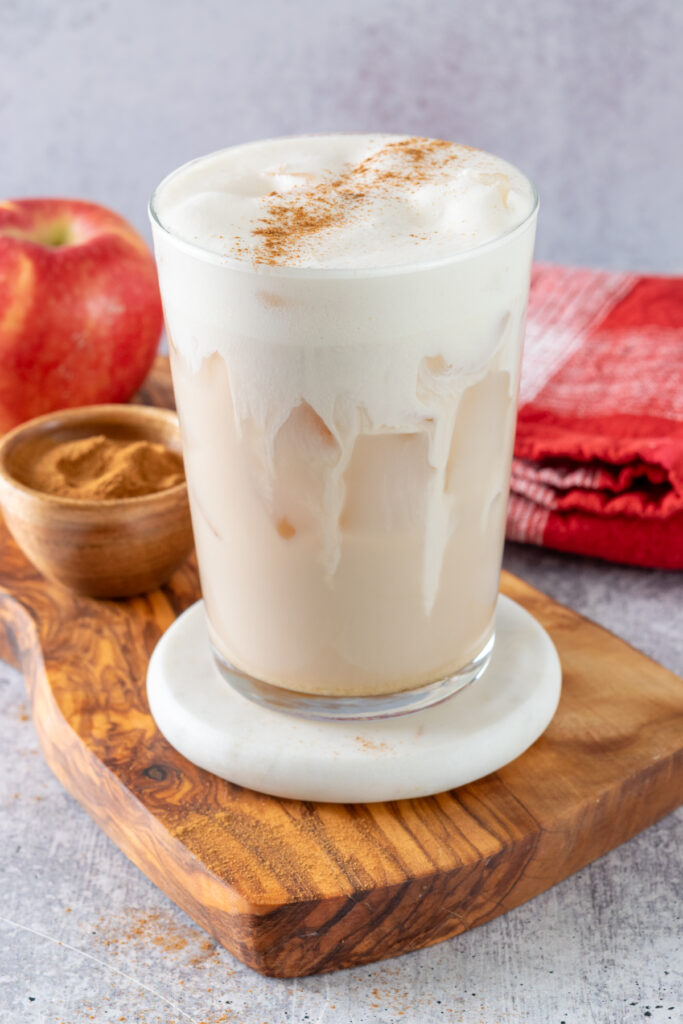 A layered, homemade iced apple crisp cream chai with cinnamon sprinkled on top. The chai drink is next to a small bowl of cinnamon and an apple.