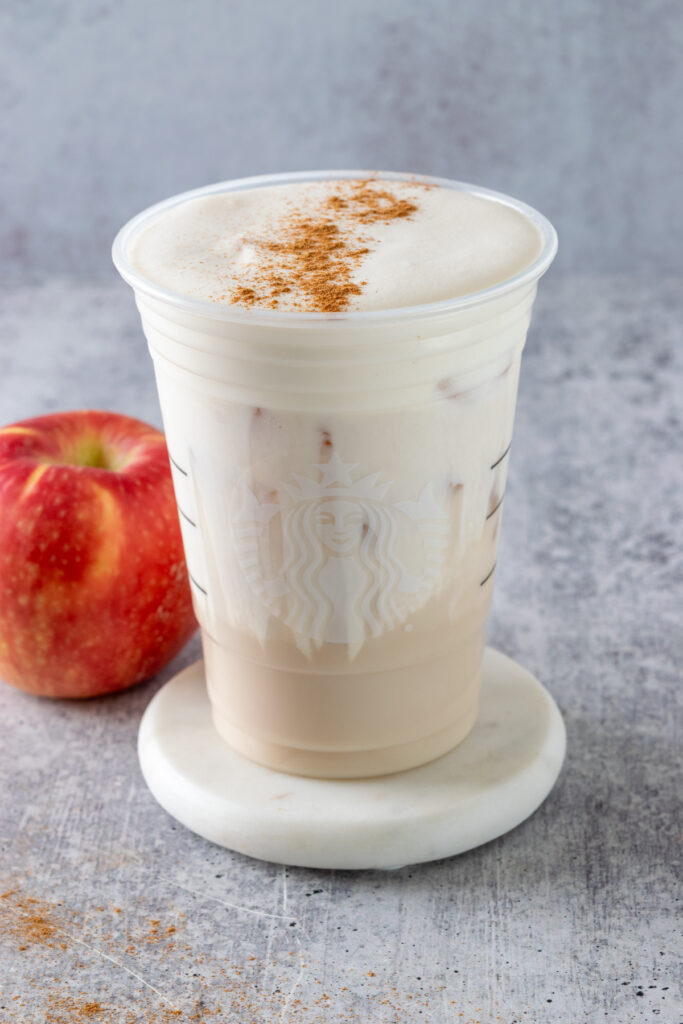 A homemade apple crisp cream chai latte in a Starbucks cup with cinnamon sprinkled on top of the creamy cold foam. An apple is sitting next to the drink which is on a white coaster.