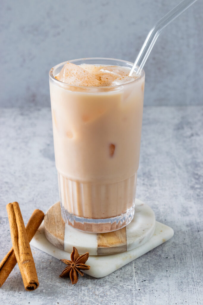 A homemade ice chai latte with cinnamon on top and a glass straw, next to two cinnamon sticks and one star anise.