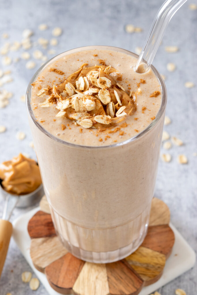 A homemade banana smoothie in a glass showing the top of the smoothie that is garnished with a few slices of banana, some oats and a little peanut butter. The smoothie has a glass straw, is on a wooden coaster and next to a spoon with a scoop of peanut butter in it.