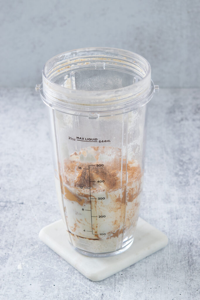 A blender cup with all the ingredients in it needed to make a banana oatmeal smoothie, including oats, frozen banana chunks, peanut butter, maple syrup, cinnamon, salt and milk.