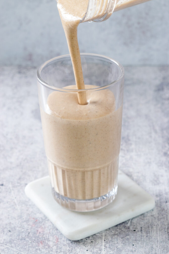 Pouring a thick and creamy, blended banana oatmeal peanut butter smoothie into a tall glass.