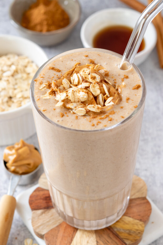 A top-down look at a delicious banana oatmeal smoothie that has banana slices, peanut butter and oats sprinkled on top. The smoothie is next to a scoop of peanut butter, bowl of oats, and small bowls with maple syrup and cinnamon.