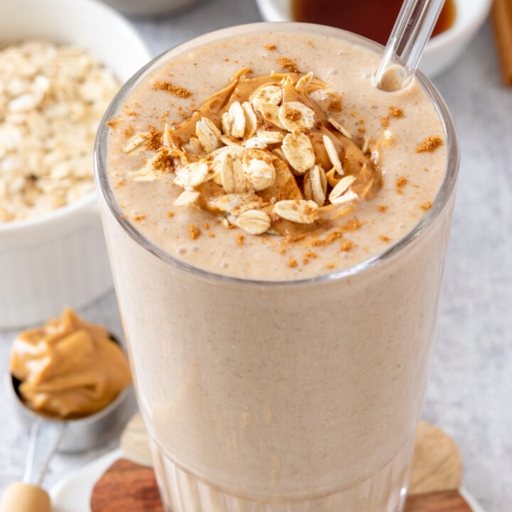 Homemade banana oatmeal smoothie with peanut butter, in a glass with a glass straw, and garnished with slices of banana, peanut butter drizzle and a few oats sprinkled on top.