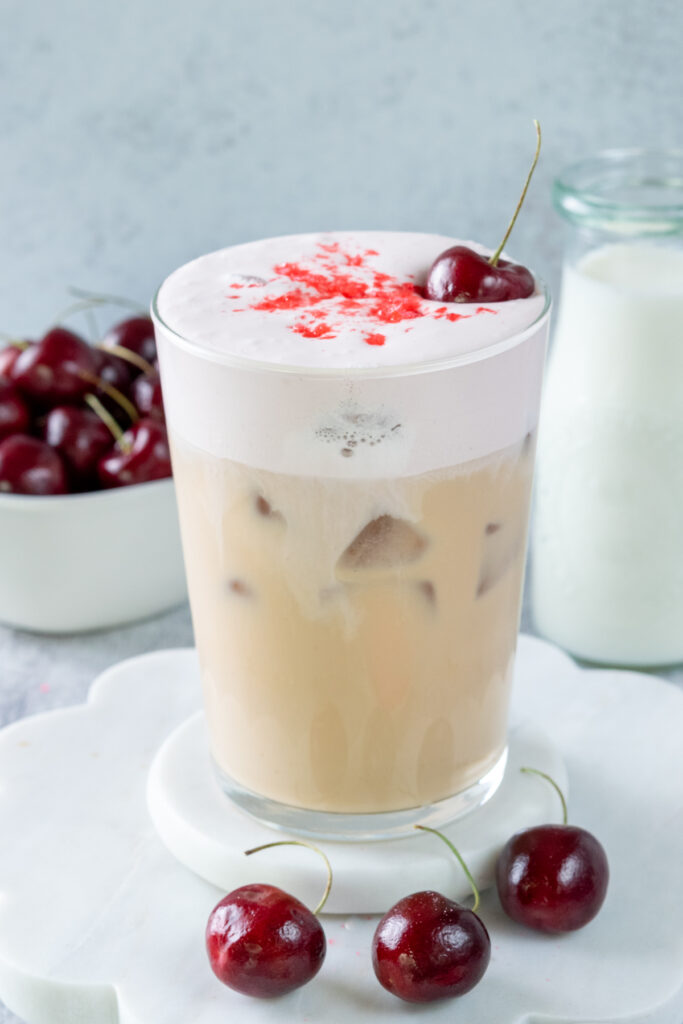 An iced cherry chai in a glass showing the layers in the drink with iced chai on the bottom and a pink colored cherry cream cold foam on top. The drink is garnished with crushed cherry sprinkles, a fresh cherry. In the background is a bowl of cherries and a pitcher of milk.