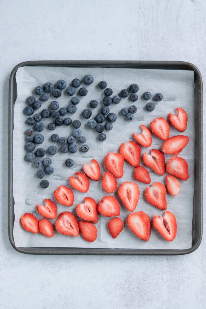 A parchment paper-lined baking tray that has a single layer of fresh berries to freeze. Half the tray is blueberries and the other half is hulled and sliced strawberries.