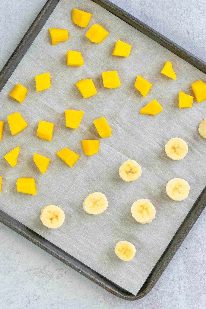 Chunks of fresh mango and banana on a tray lined with parchment paper, ready to be placed in freezer.