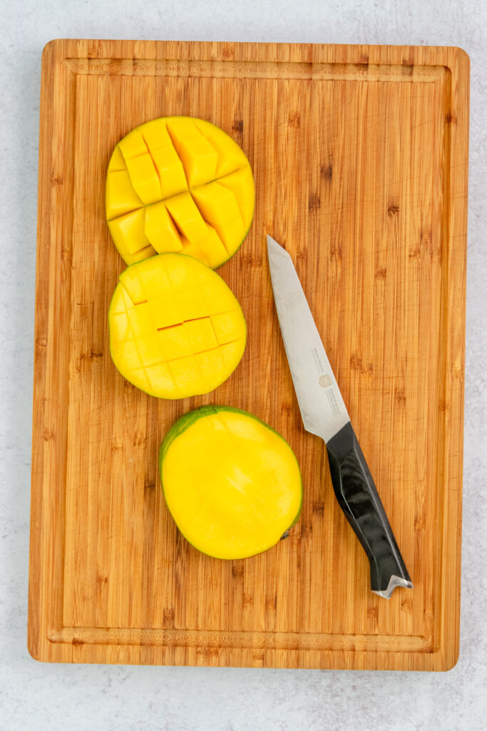 A fresh mango on a cutting board next to a knife that's being cut into small chunks.