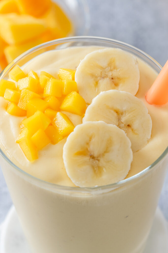 A close-up view of the top of a mango banana smoothie that has three slices of banana and some chopped, fresh mango on top. The smoothie has an orange straw in it and you can see how thick and creamy it is.