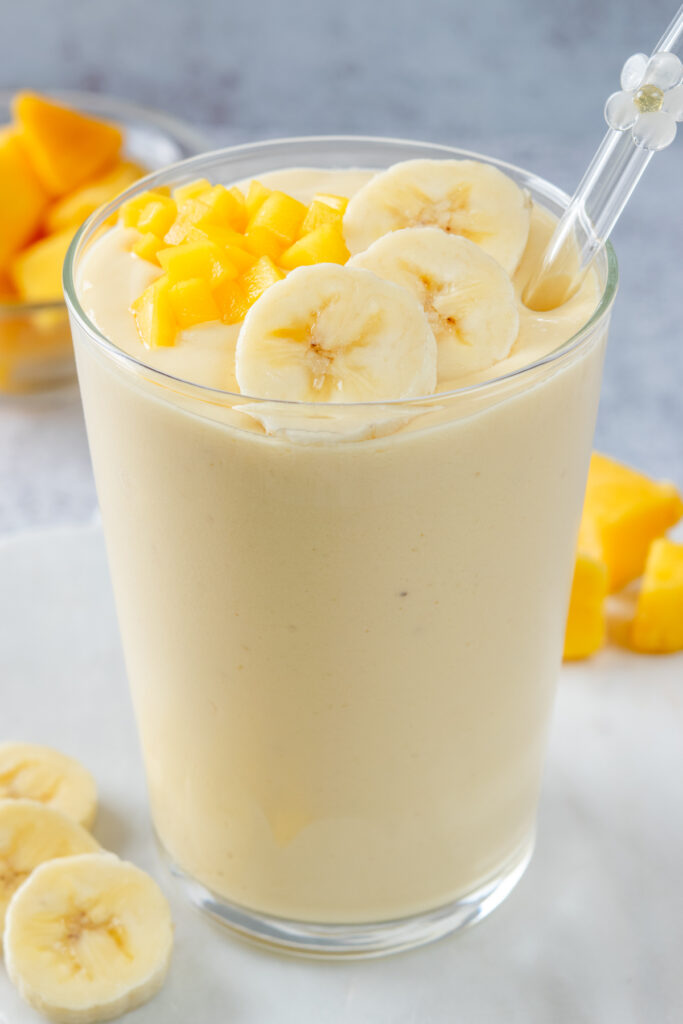 A super creamy, homemade mango banana smoothie in a glass cup that has a glass straw in it. The straw has a little white glass flower on it, and the cup is next to a bowl of mango chunks  and a sliced banana.