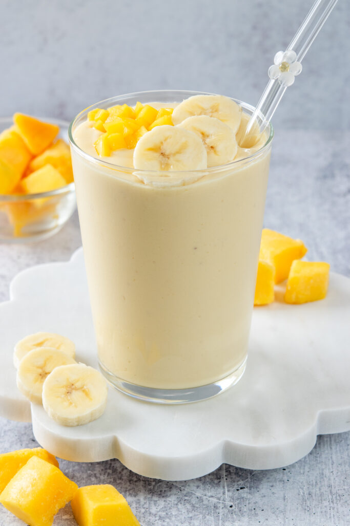 A homemade mango banana smoothie garnished with a few banana slices and chopped fresh mango. The smoothie is in a glass with a glass straw that has a white flower on it.